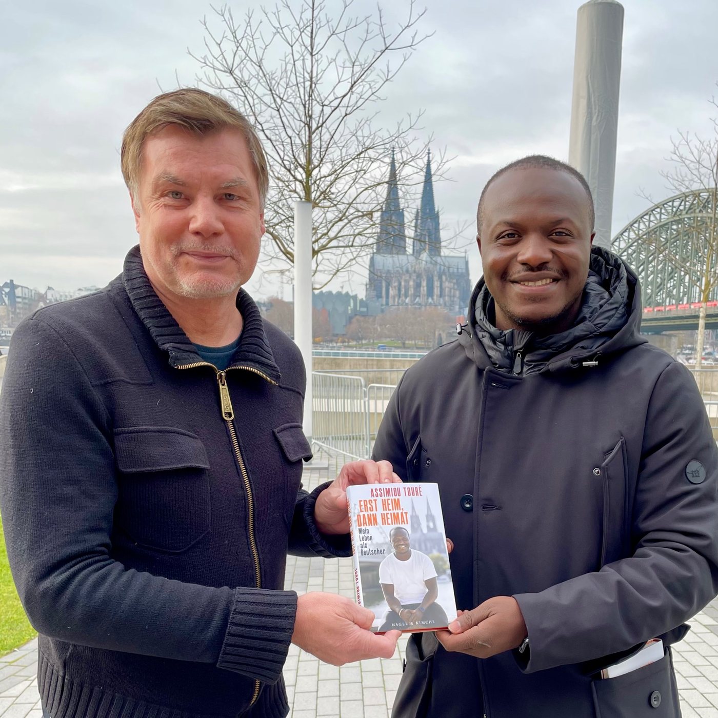 Zwei Fußball-Profis am Kölner Rheinufer: Thomas Helmer und Assimiou Touré halten gemeinsam das Buch „Erst Heim, dann Heimat“, im Hintergrund der Kölner Dom